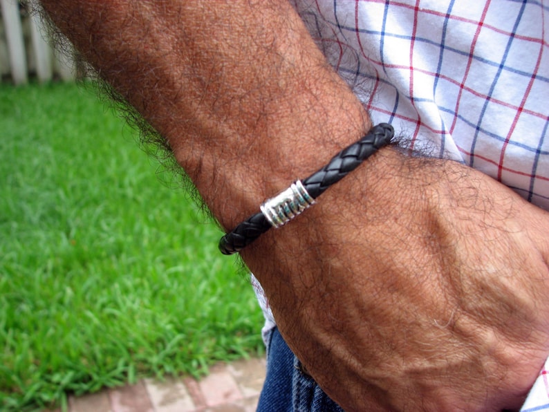 May include: A black leather bracelet with a silver metal clasp. The bracelet is worn on a person's wrist.