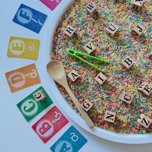 Puede incluir: Un juego de actividades sensoriales con un cuenco blanco lleno de fideos de colores y bloques de letras de madera. También se incluyen tarjetas del alfabeto, una cuchara de madera y pinzas verdes. Juguete educativo.