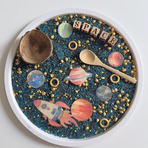 Puede incluir: Una bandeja de juego sensorial con temática espacial. La bandeja contiene arroz azul y dorado, planetas de madera, un cohete, una cuchara de madera y la palabra "SPACE" escrita con bloques de madera.