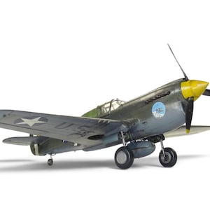May include: A scale model of a P-40 Warhawk fighter aircraft featuring a dark green and grey camouflage paint scheme, a bright yellow propeller spinner, a clear cockpit canopy, and a white star insignia on the wing against a plain white background.