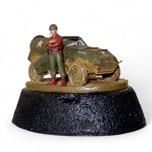 Może przedstawiać: Miniaturowy model wojskowego pojazdu zwiadowczego w kolorze oliwkowym z napisem JEANIE oraz tekstem 815-6 na boku. Figurka żołnierza w hełmie, czerwonej koszuli i zielonych spodniach stoi obok pojazdu, całość umieszczona na czarnej podstawie.