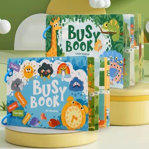 May include: Two children's "Busy Book" activity books. One book features a giraffe and the text "Look! Animal". The other book has a clock and the text "Hi! Weather". The books have colorful illustrations and are bound with blue rings.