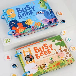 May include: Two colorful Busy Books with ring binding. One features sea creatures and the text "Yeah! Learning Maths." The other has farm animals and the text "Hello! Alphabet!" Several number tiles are scattered around the books.