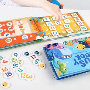 May include: A colorful educational toy set featuring a book with letters and illustrations, a book with a whale and fish, and number tiles. The set is designed for learning and play, with bright colors and engaging graphics.