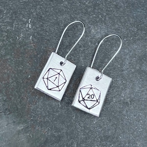 May include: A pair of silver earrings with a rectangular charm featuring a black and white outline of a twenty-sided die with the number "20" on one earring and the number "1" on the other earring.