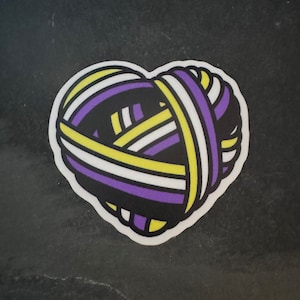 May include: A heart-shaped yarn ball with purple, yellow, and white stripes. The yarn is tied together in a knot at the top.