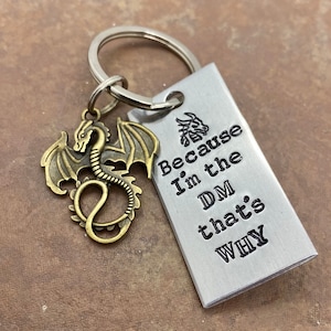 May include: A silver keychain with a dragon charm and a silver tag that reads "Because I'm the DM that's WHY".