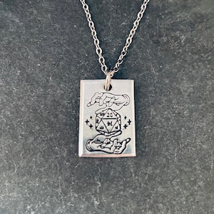 May include: Silver necklace with a rectangular pendant featuring two hands holding a 20-sided die with stars around it.