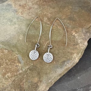 May include: A pair of silver-toned earrings featuring long, curved ear wires and small, circular charms. Each charm is embossed with a repeating, textured herringbone or knit-like pattern across the entire surface of the metal disc.