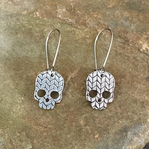 May include: A pair of silver-toned drop earrings featuring a skull-shaped pendant. The surface of each skull is embossed with a textured pattern resembling a knitted stitch, and each piece hangs from a simple silver-toned wire hook.