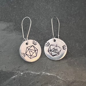 May include: A pair of silver earrings with a 20-sided die and a 1-sided die engraved on the front. Each die is surrounded by handprint designs.
