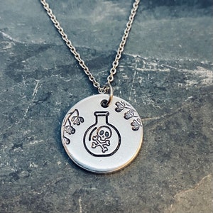 May include: Silver necklace with a round pendant featuring a skull and crossbones inside a vial, surrounded by floral designs.