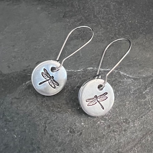 May include: A pair of silver dangle earrings with a dragonfly design. The earrings are made of metal and have a simple, elegant design.