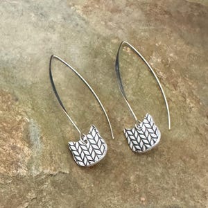 May include: A pair of silver-toned earrings featuring a cat-shaped charm with a textured, herringbone-like pattern. The earrings are suspended from long, curved, open-loop ear wires that extend upward from the top of each cat-shaped pendant.