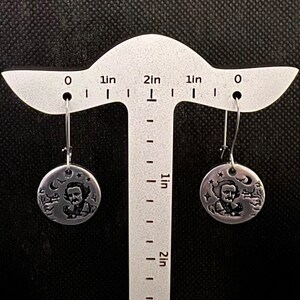 May include: A pair of silver dangle earrings featuring a round disc with an image of Edgar Allan Poe and a raven. The disc is approximately 1 inch in diameter.
