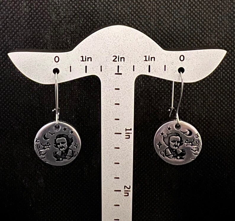 May include: A pair of silver dangle earrings featuring a round disc with an image of Edgar Allan Poe and a raven. The disc is approximately 1 inch in diameter.