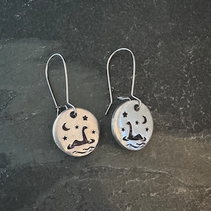May include: A pair of silver earrings with a circular design featuring a silhouette of a Loch Ness Monster in a lake with stars and a crescent moon.