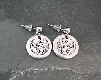 May include: A pair of silver earrings with a circular design featuring a black and white otter holding a fish. The earrings have a simple post design.