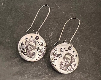 May include: Silver earrings with a round pendant featuring a black and white illustration of a man with a beard and a crescent moon. The pendant is surrounded by stars and a tree branch.
