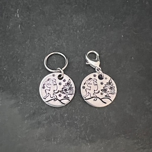 May include: Two silver keychains with a winter scene design. One keychain features a snowman and a Christmas tree, while the other keychain features a reindeer and a Christmas tree. Both keychains have a silver ring.