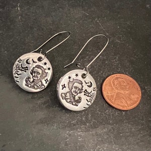 May include: A pair of silver earrings featuring a round disc with an engraved image of Edgar Allan Poe, a crescent moon, stars, and a raven. The earrings are approximately the size of a US penny.