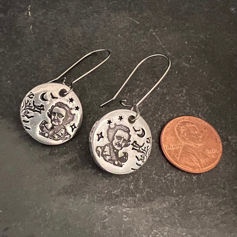 May include: A pair of silver earrings featuring a round disc with an engraved image of Edgar Allan Poe, a crescent moon, stars, and a raven. The earrings are approximately the size of a US penny.