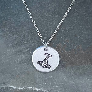 May include: A silver necklace with a round pendant featuring a detailed engraved image of a hammer with intricate designs.