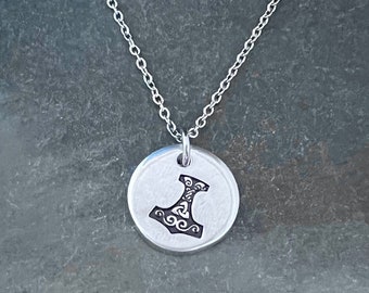 May include: A silver necklace with a round pendant featuring a detailed engraved image of a hammer with intricate designs.