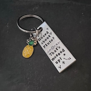 May include: A silver keychain with a pineapple charm and a rectangular tag that reads "Heard about Pluto? That's messed up!" with a star and a constellation design.