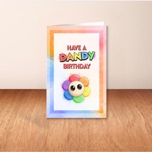 May include: A folded greeting card with a watercolor-style border in shades of orange, blue, and purple. The card features the text "HAVE A DANDY BIRTHDAY" above a cartoon flower with a smiling face and colorful petals.