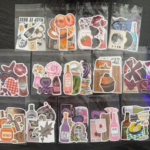 Può includere: Una collezione di pacchetti di adesivi con vari disegni. Gli adesivi includono immagini di cibo, bevande, fiori e testo. Alcuni pacchetti hanno un tema vinicolo, mentre altri presentano una varietà di illustrazioni e frasi.