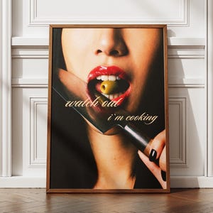 May include: A framed art print featuring a close-up of a person's mouth with red lipstick, holding a spoon with a green olive. The text "watch out i'm cooking" is displayed on the print. The frame is brown.