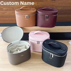 May include: Five personalised cosmetic bags in shades of brown, burgundy, pink, grey, and black. Each bag features a silver zip, a top handle, and the name "Lisa" embossed. The text "Custom Name" is at the top.