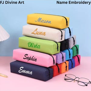 Op de afbeelding: Een stapel gepersonaliseerde etuis in diverse kleuren, waaronder geel, crème, groen, roze en marineblauw. Elke etui heeft een geborduurde naam in een contrasterende kleur en een zwarte ritssluiting.