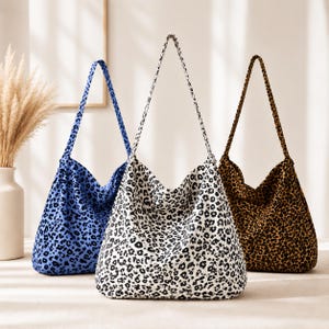 Pode incluir: Três bolsas tipo hobo com estampa de leopardo. A bolsa à esquerda é azul e preta, a bolsa central é branca e preta e a bolsa à direita é marrom e preta. Cada bolsa possui uma alça de ombro única e uma silhueta descontraída.