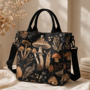 May include: A black tote bag featuring a detailed botanical print of various mushrooms, leaves, and berries in shades of brown, tan, and cream. The bag includes two black top handles, a detachable black shoulder strap, and a silver snap closure.