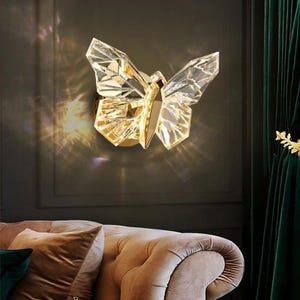 May include: A crystal butterfly wall sconce with a gold base. The butterfly's wings are made of faceted crystal, reflecting light. The sconce is mounted on a dark wall, with a sofa and green curtain in the background.