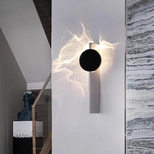May include: Modern wall sconce with a black circular shade and a rectangular silver base. The light casts a unique pattern on the wall. The sconce is mounted on a white wall, adding a contemporary touch to the interior decor.
