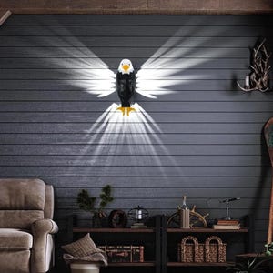May include: A wall-mounted eagle sculpture with outstretched wings, featuring a black body, white wings, and yellow talons. The sculpture casts a dramatic light pattern on the dark gray wooden wall, creating a striking visual effect. The eagle is a decorative element.