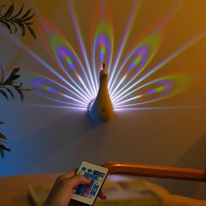 Op de afbeelding: Een wandlamp in de vorm van een pauw die een kleurrijk, waaierachtig lichtpatroon projecteert. De lamp is lichtgeel en is bevestigd aan een lichtgekleurde muur. Een afstandsbediening is zichtbaar op de voorgrond.