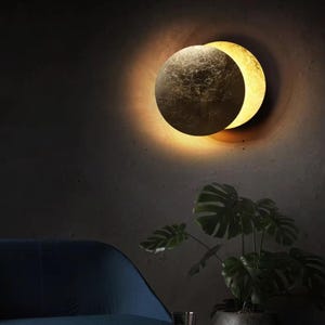 Op de afbeelding: Een moderne wandlamp met een cirkelvormig ontwerp en een halve maanvorm. De lamp heeft een getextureerde, donkere metalen afwerking met een warme, gele gloed. Een kamerplant staat op de voorgrond, met een blauwe stoel linksonder.