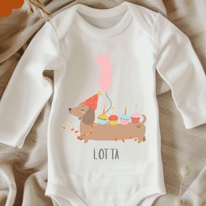 Op de afbeelding: Witte babybody met lange mouwen met een illustratie van een teckel met een feesthoed, cupcakes en een roze ballon. De tekst "LOTTA" staat eronder.