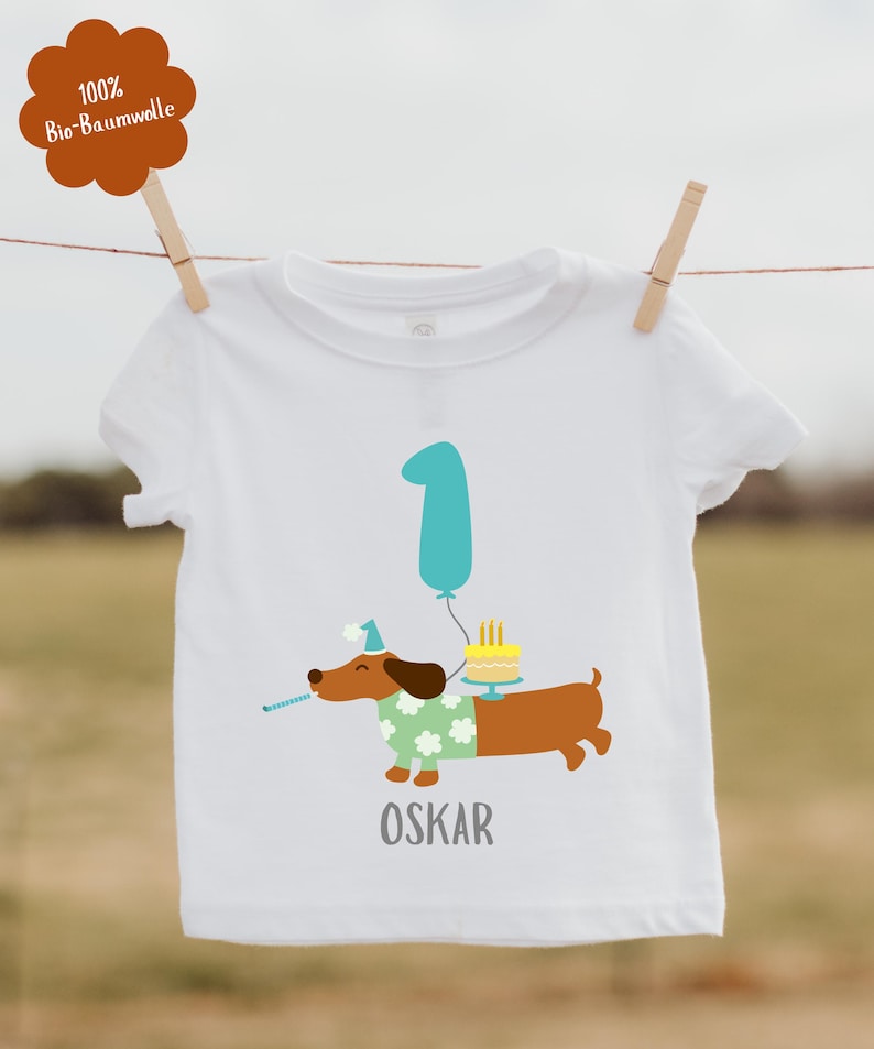 Pu&ograve; includere: T-shirt bianca a maniche corte con un bassotto dei cartoni animati con cappello e maglione da festa, che tiene un corno da festa e porta una torta. Un palloncino turchese con il numero 1 e il nome "OSKAR" sono stampati. 100% cotone biologico.