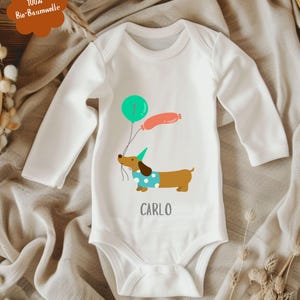 Op de afbeelding: Witte babybody met lange mouwen. Het heeft een cartoon teckel met ballonnen, een groene ballon met het nummer &eacute;&eacute;n en een roze worstvormige ballon. De naam "CARLO" staat eronder gedrukt. De body is gemaakt van 100% biologisch katoen.