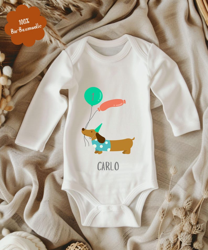 Op de afbeelding: Witte babybody met lange mouwen. Het heeft een cartoon teckel met ballonnen, een groene ballon met het nummer &eacute;&eacute;n en een roze worstvormige ballon. De naam "CARLO" staat eronder gedrukt. De body is gemaakt van 100% biologisch katoen.