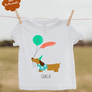 Pu&ograve; includere: Maglietta bianca per bambini con un bassotto dei cartoni animati che tiene dei palloncini. Il palloncino &egrave; verde e arancione. Il nome "CARLO" &egrave; stampato sotto. Un'etichetta marrone con testo &egrave; attaccata alla maglietta.