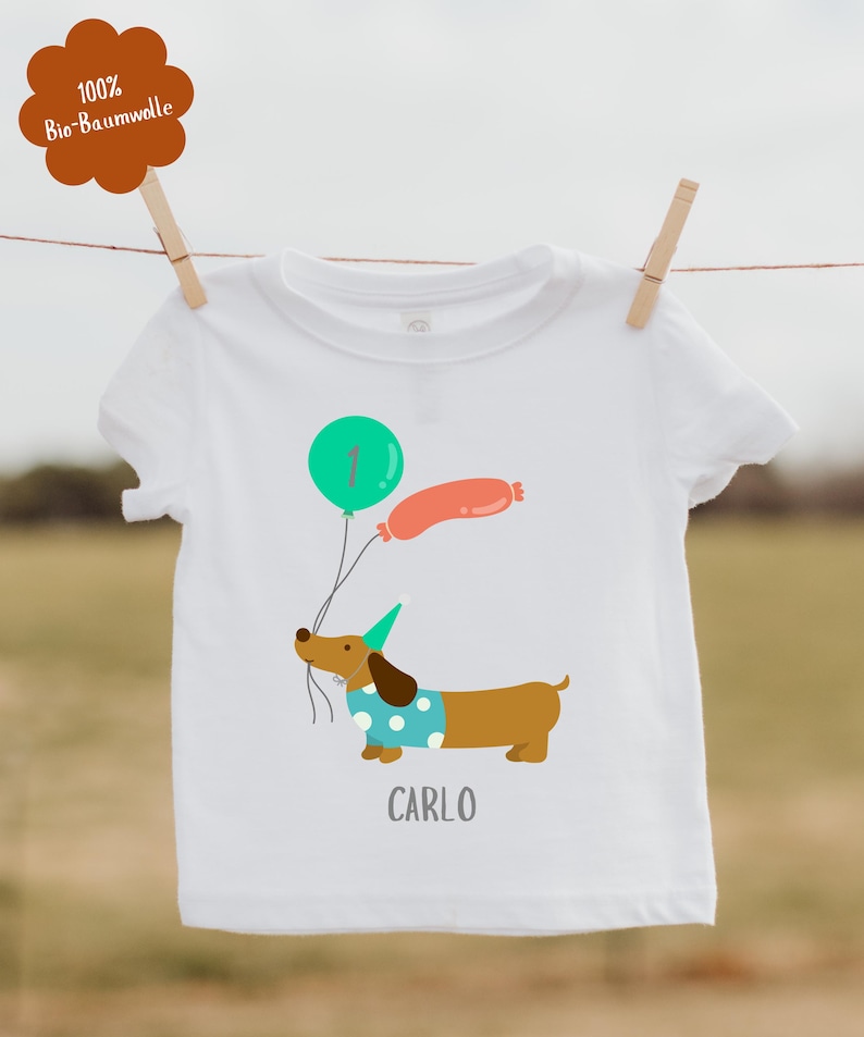 Pu&ograve; includere: Maglietta bianca per bambini con un bassotto dei cartoni animati che tiene dei palloncini. Il palloncino &egrave; verde e arancione. Il nome "CARLO" &egrave; stampato sotto. Un'etichetta marrone con testo &egrave; attaccata alla maglietta.