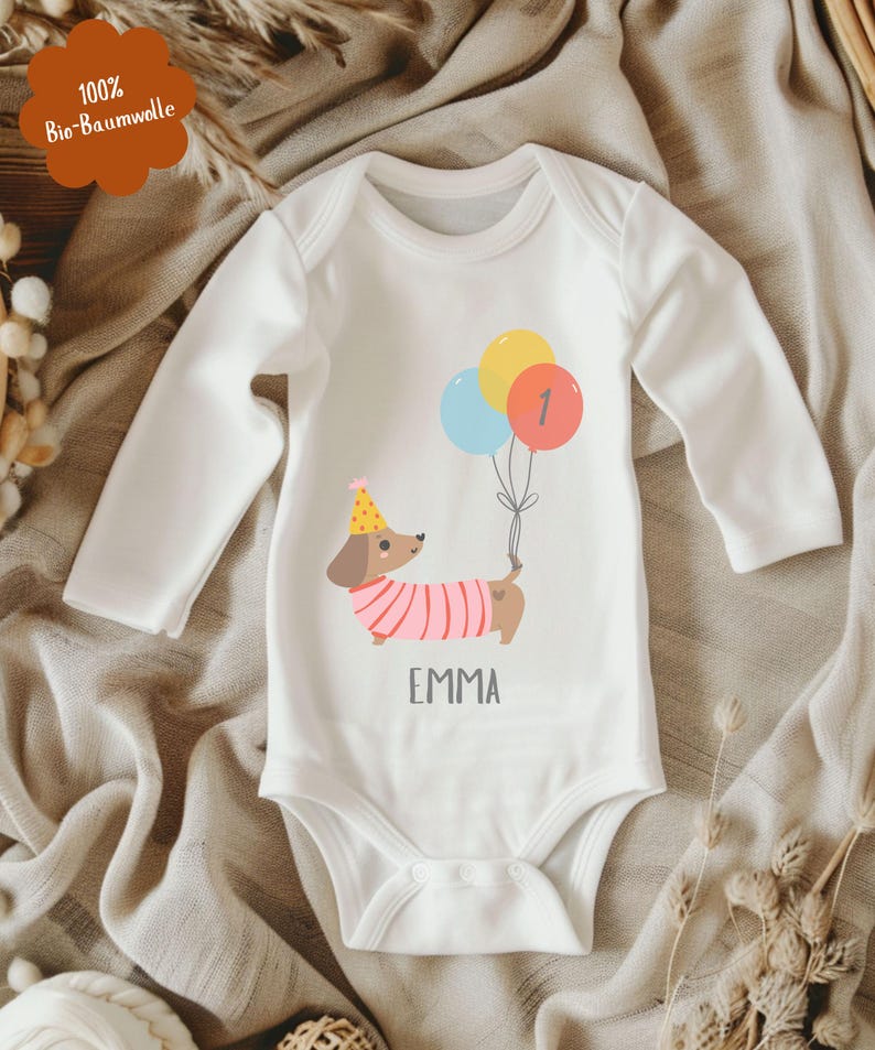 Op de afbeelding: Witte babybody met lange mouwen. De body heeft een cartoon teckel met een feesthoed en een roze gestreepte trui, die ballonnen vasthoudt. De ballonnen zijn geel, blauw en rood met het nummer 1. De naam "EMMA" staat eronder gedrukt.