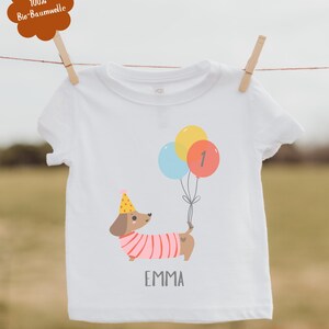 Pu&ograve; includere: T-shirt bianca con un bassotto dei cartoni animati con un cappello da festa e palloncini. I palloncini sono gialli, blu e rossi con il numero 1. Il nome "EMMA" &egrave; stampato sotto.