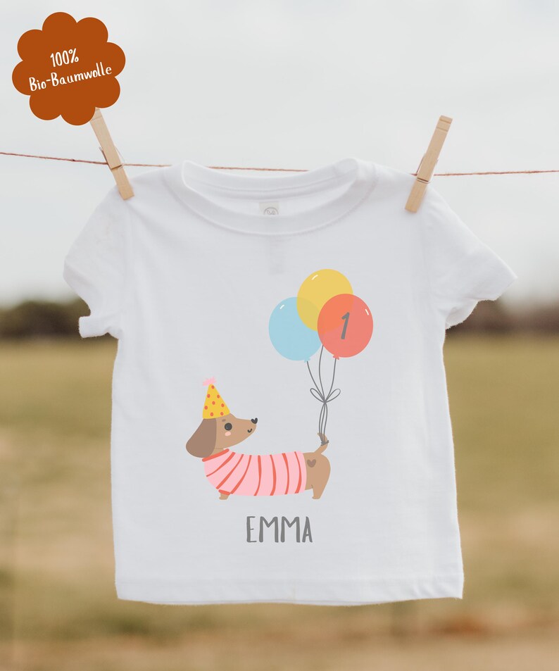 Pu&ograve; includere: T-shirt bianca con un bassotto dei cartoni animati con un cappello da festa e palloncini. I palloncini sono gialli, blu e rossi con il numero 1. Il nome "EMMA" &egrave; stampato sotto.